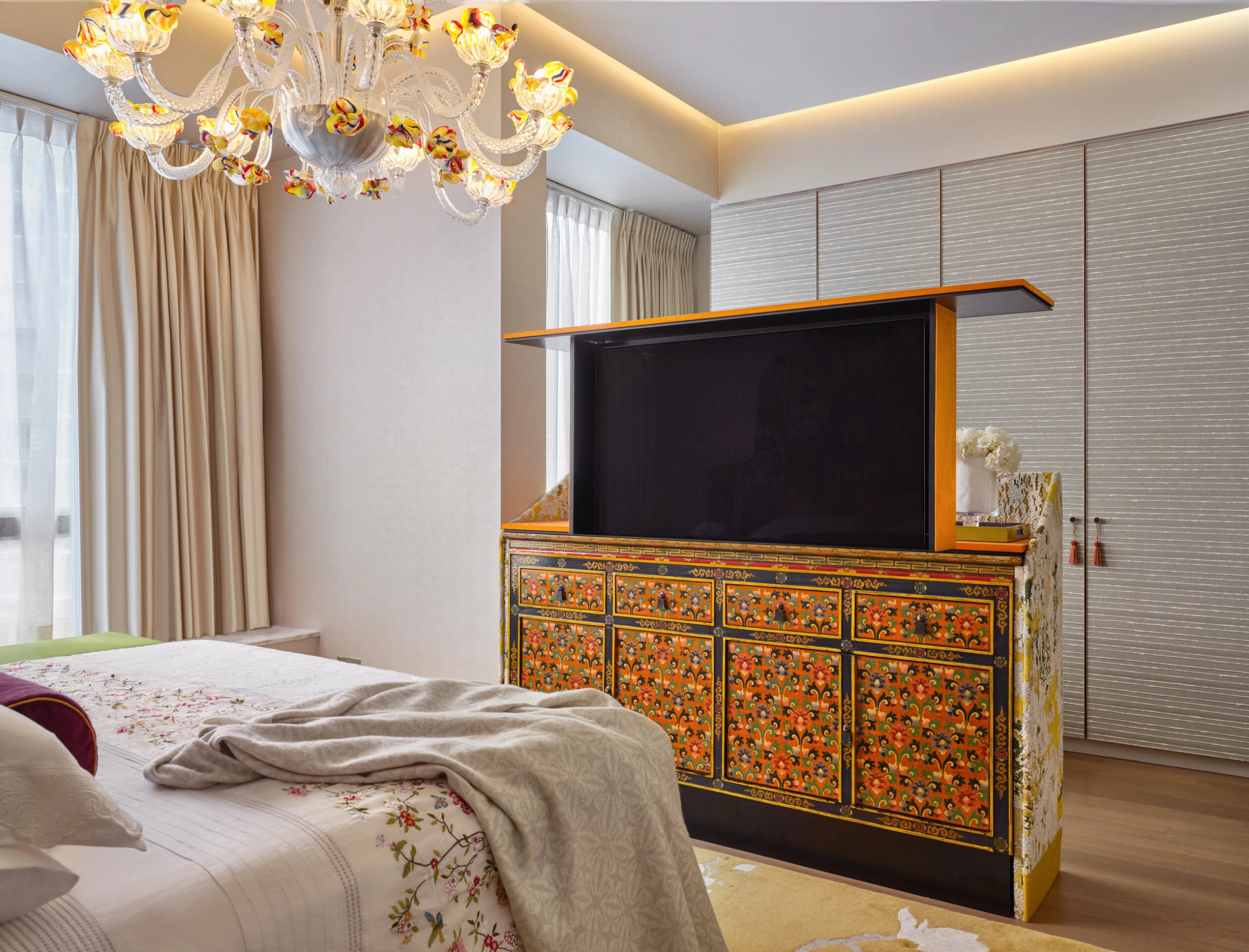 Luxury bedroom with ornate floral-painted cabinet housing a flat-screen TV, Murano glass chandelier, and neutral bedding with embroidered accents.