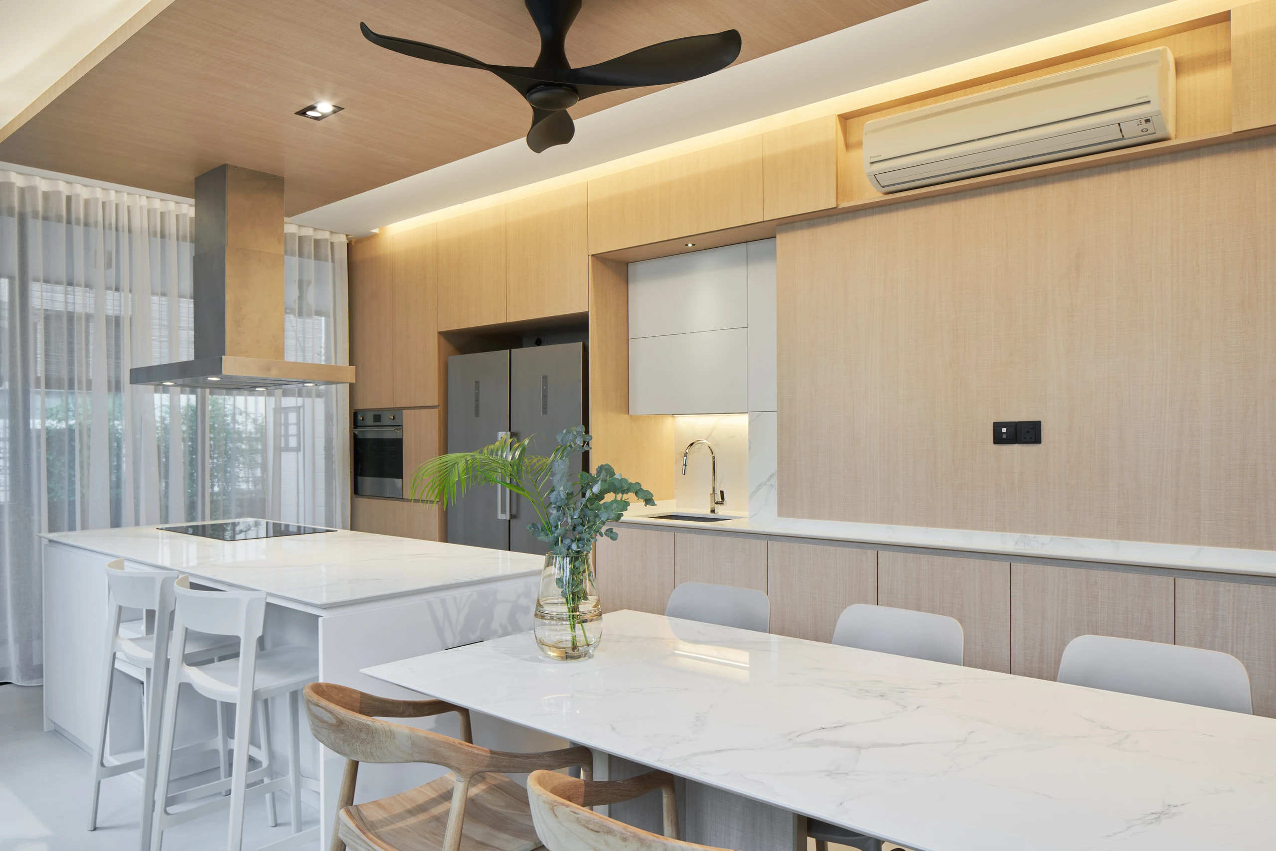 Open-plan kitchen and dining area with white marble table, light wood cabinetry, ceiling fan, and sheer curtains
