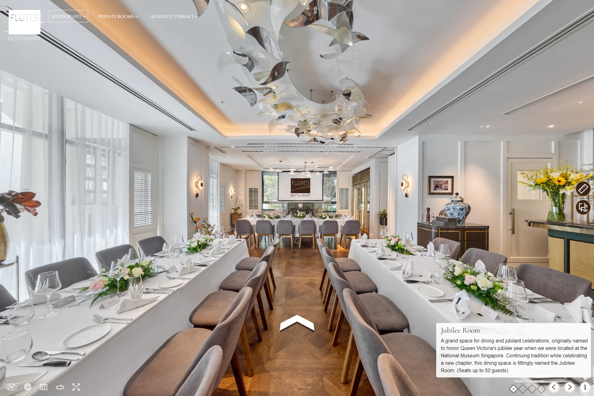Virtual tour panoramic view of a restaurant private dining room with long white-clothed tables, floral centerpieces, grey chairs, and a metallic leaf ceiling sculpture.