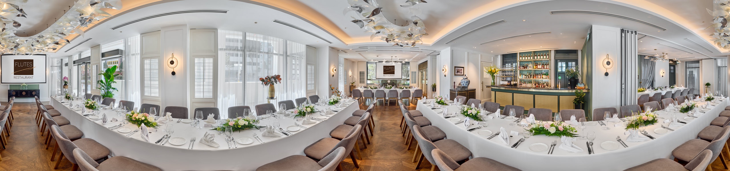 Panoramic view of a white-tablecloth restaurant with banquet seating, floral centrepieces, grey upholstered chairs, a bar area, and sculptural ceiling art.
