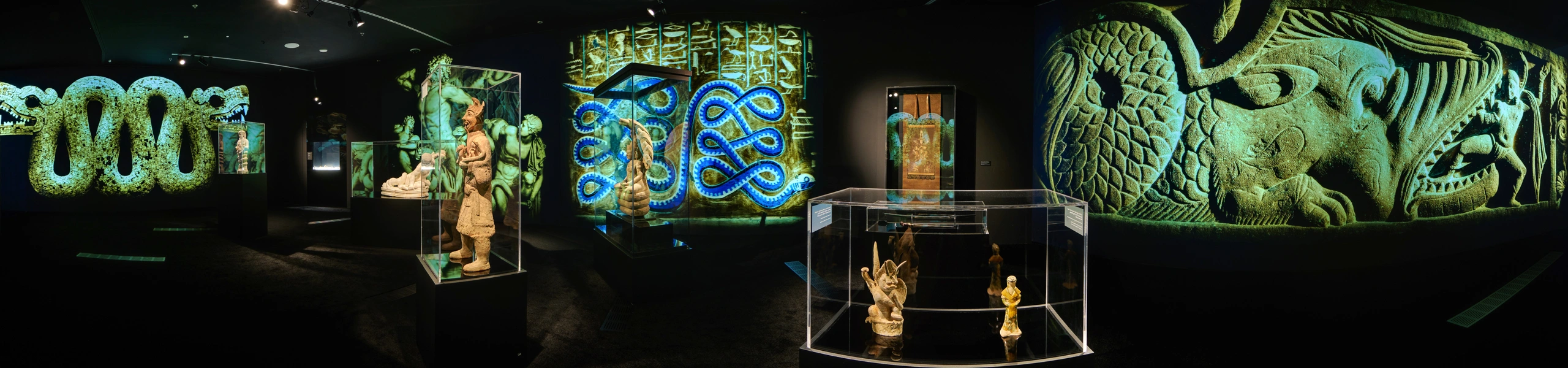 Panoramic view of museum gallery with ancient serpent sculptures in glass cases, intertwined snake wall projections, and stone relief imagery