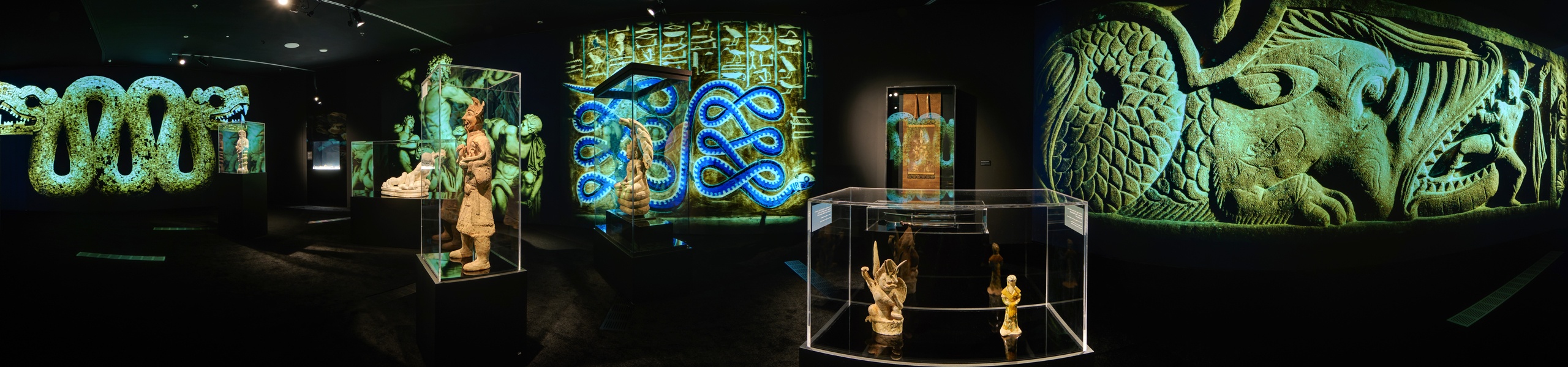Panoramic view of museum gallery with ancient serpent sculptures in glass cases, intertwined snake wall projections, and stone relief imagery