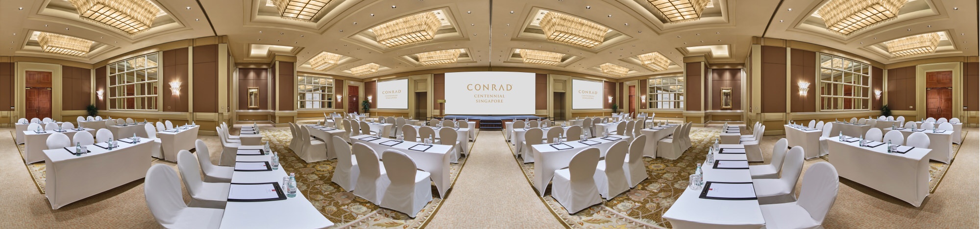 Panoramic view of a grand hotel ballroom in classroom setup with white-draped tables and chairs, central projection screen, coffered gold ceiling, and mirrored wall panels.