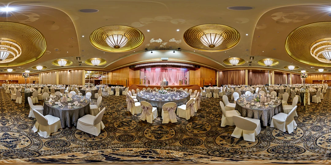 Grand ballroom set for a banquet with round tables draped in white and grey, sashed chairs, large gold concentric-ring chandeliers, and a pink-lit stage.