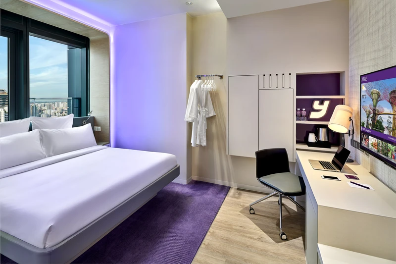 Compact hotel suite with white bedding, purple LED perimeter lighting, purple carpet, open wardrobe, long desk with laptop, and city skyline views.
