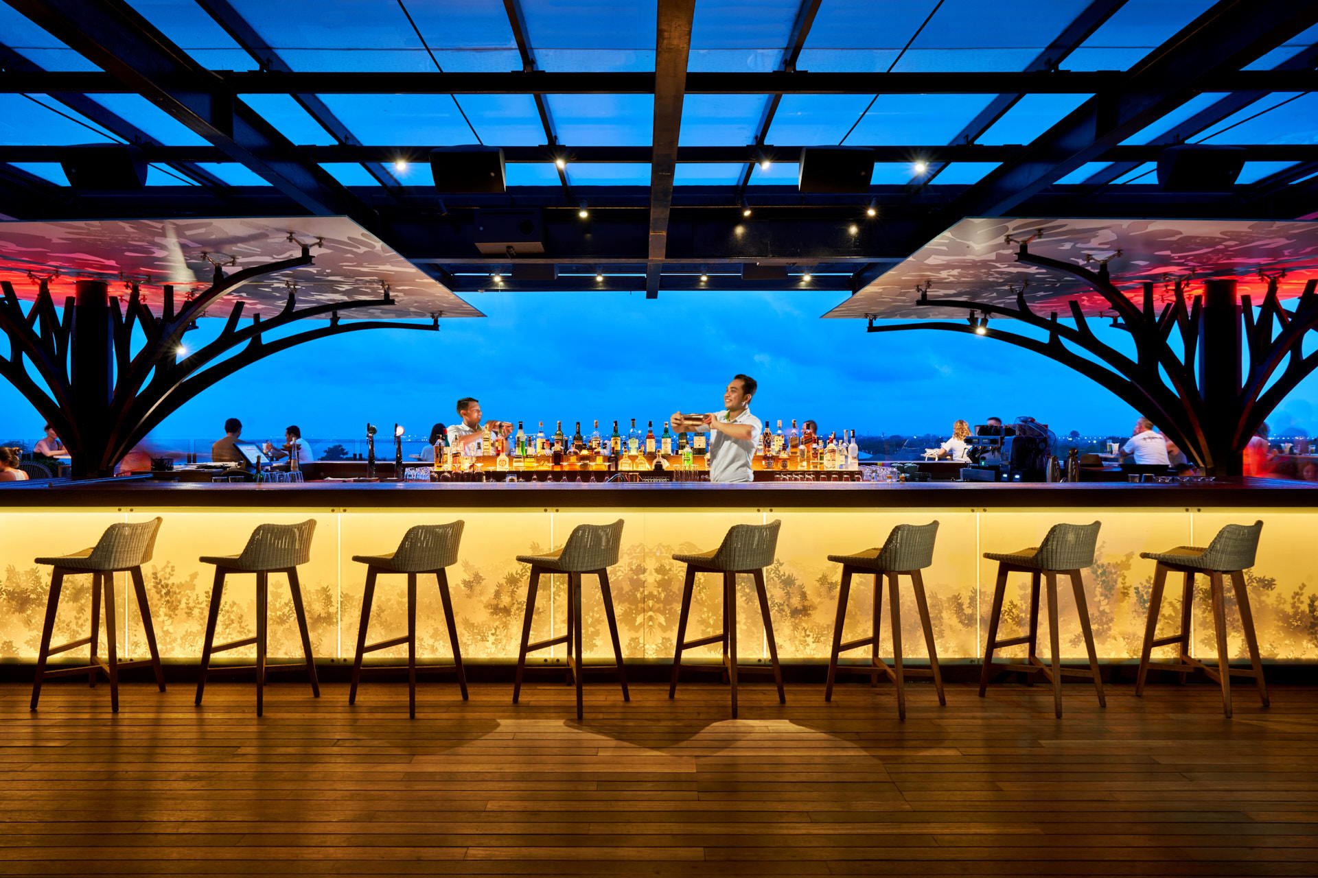 Bar at blue hour with bartender, woven stools and tropical foliage panel