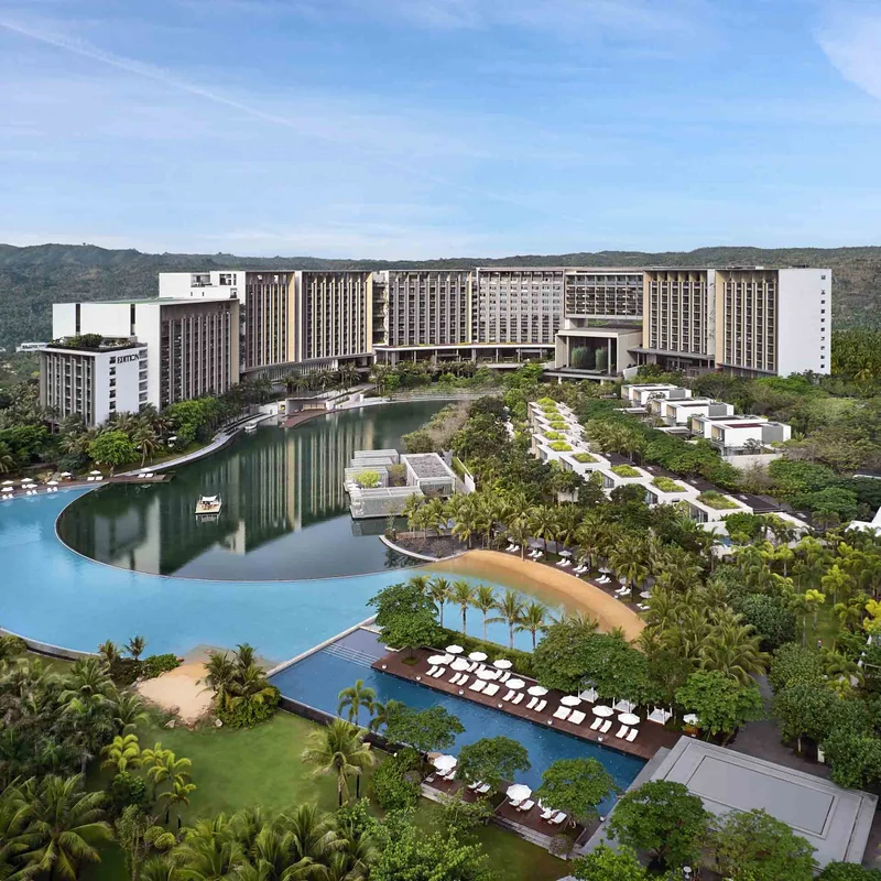 Aerial view of a modern resort hotel with a sweeping blue lagoon pool, sandy beach area, tropical landscaping, and forested hills behind.