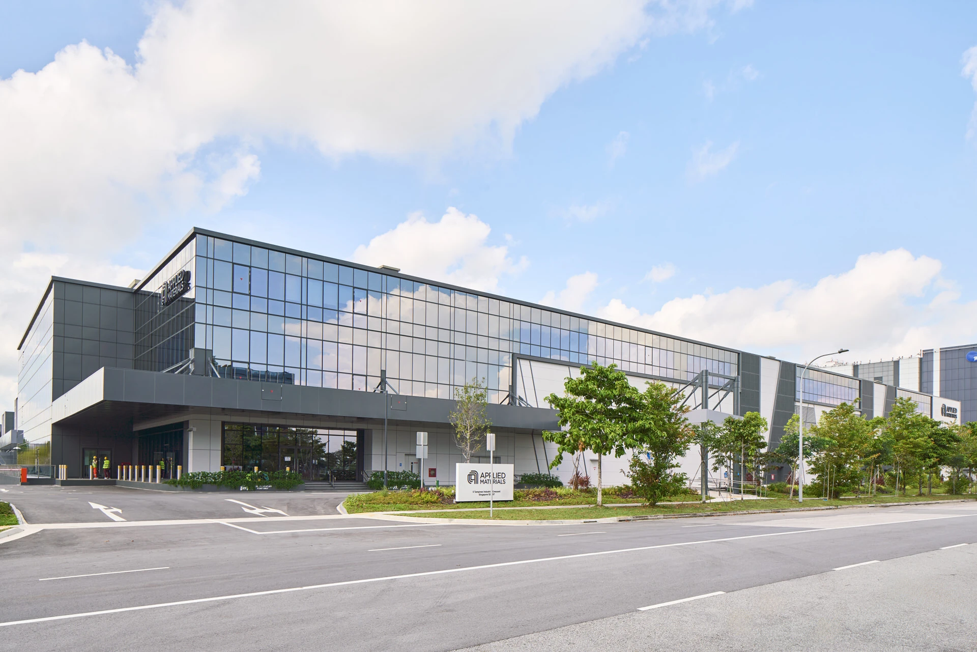 Modern industrial building with a dark glass and grey metal-clad facade, ground-level entrance canopy, landscaped trees, viewed from the roadside.