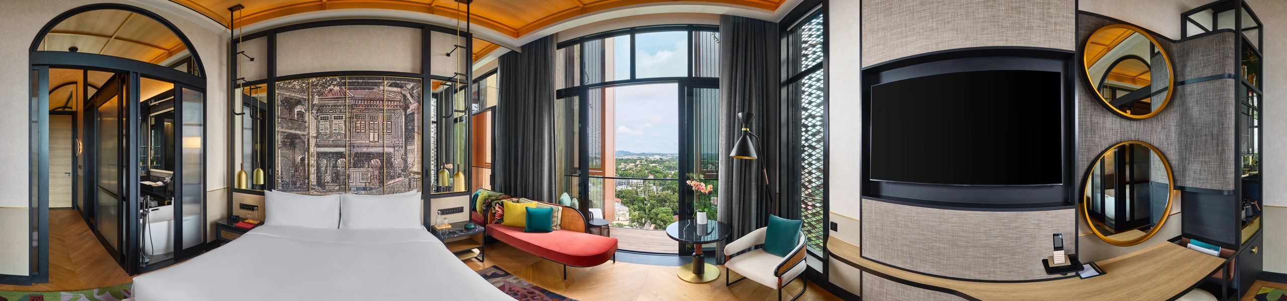 Panoramic view of a boutique hotel bedroom with king bed, shophouse mural headboard, red daybed, arched glass bathroom partition, balcony windows, and wall-mounted TV.
