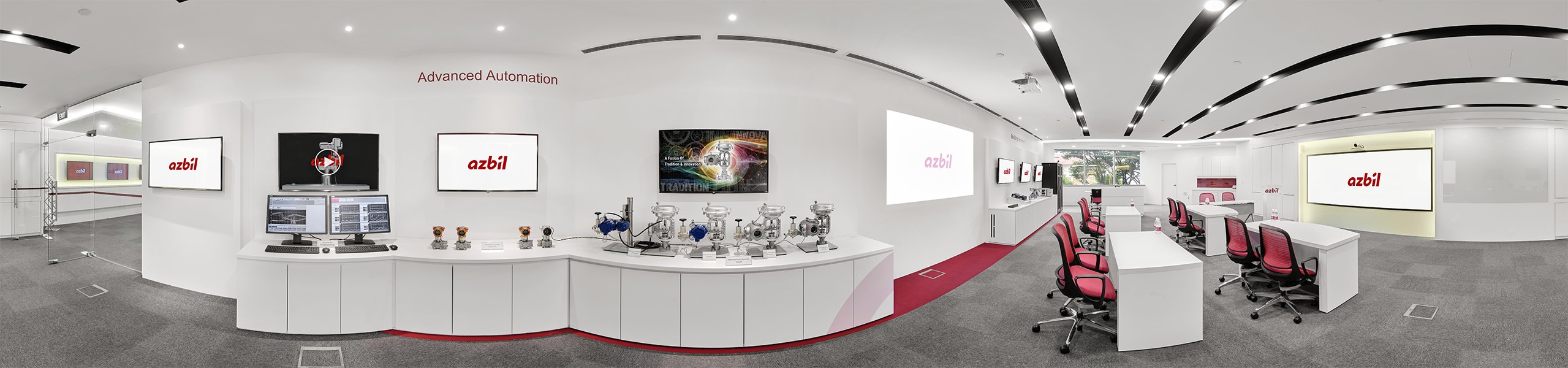 Panoramic view of a white corporate showroom displaying industrial automation valves and sensors on curved counters, with branded screens and red office chairs.