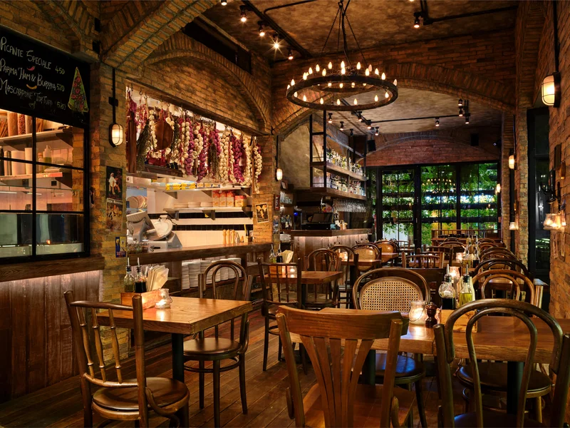 Rustic Italian restaurant interior with exposed brick arches, hanging cured meats and garlic, circular candle chandelier, bentwood chairs, and warm candlelit tables at night.