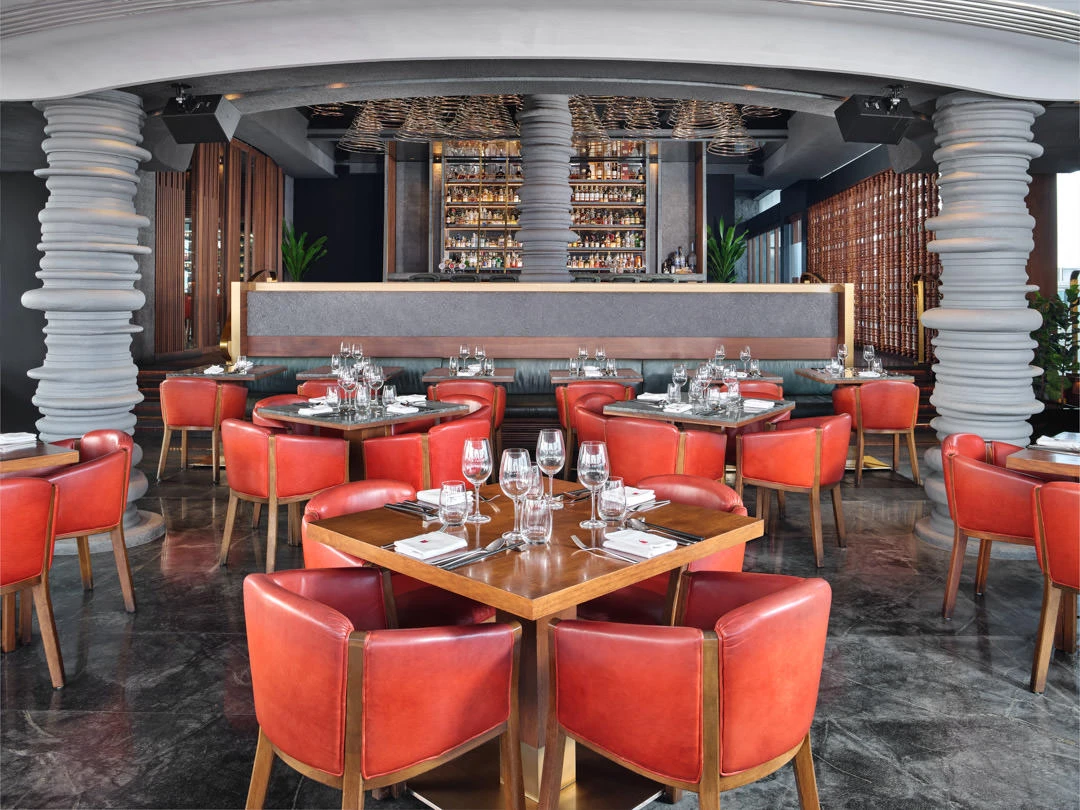 Restaurant dining room with red leather chairs, sculpted grey columns, marble floors, and backlit bar shelving