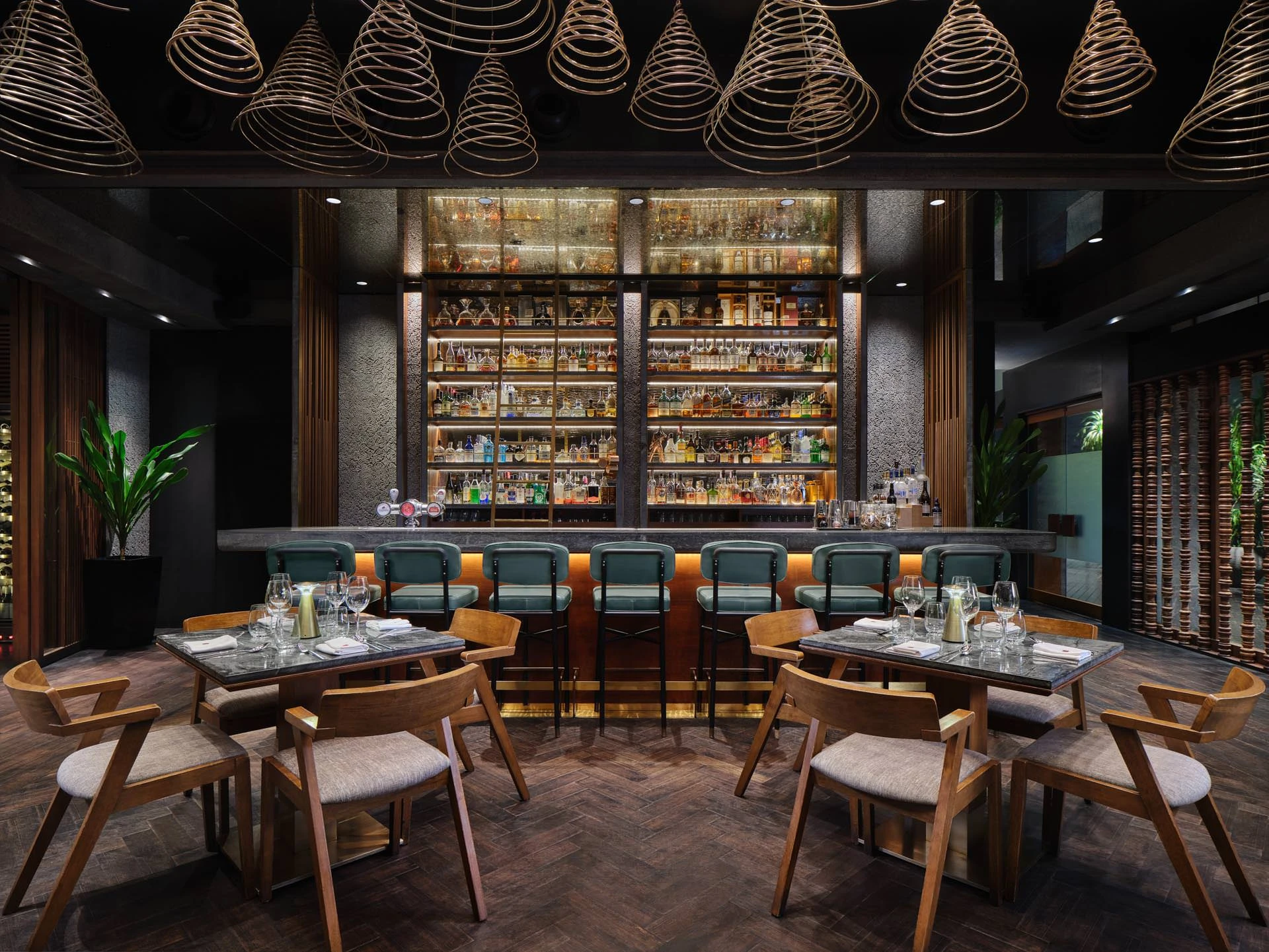 Dining area with mid-century wooden chairs and dark tables facing the main bar wall of illuminated spirit bottles behind spiral cone ceiling pendants at CÉ LA VI Singapore