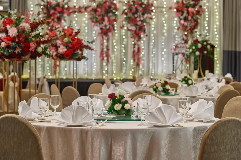 Wedding banquet ballroom with round tables set with white linens and folded napkins, low rose centerpieces, tall red floral arrangements on gold stands, and a fairy light curtain backdrop.