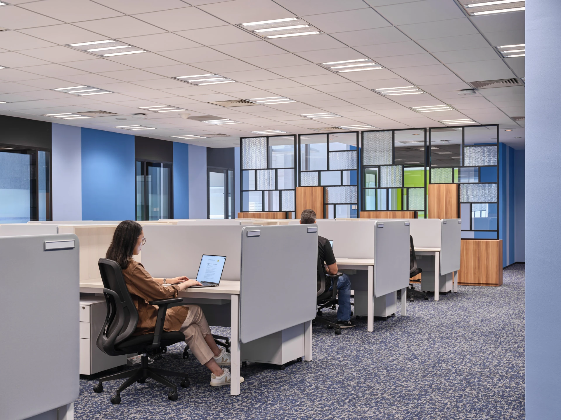Open-plan office with grey partitioned desks, two workers at laptops, blue carpet, blue and lavender accent walls, and decorative grid-panel partitions.