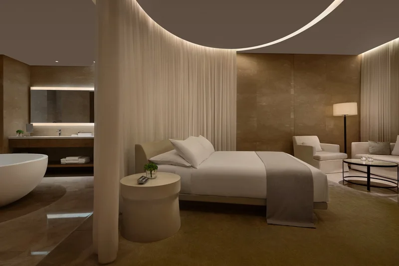 Luxury hotel suite with white bed, curved sheer curtains, freestanding bathtub and warm ambient lighting