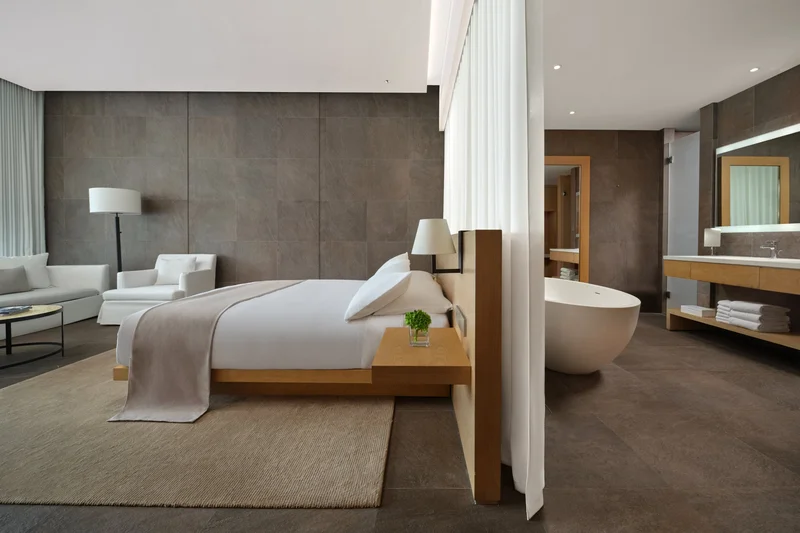 Modern open-plan hotel room with oak bed frame, freestanding white bathtub and stone-tiled walls