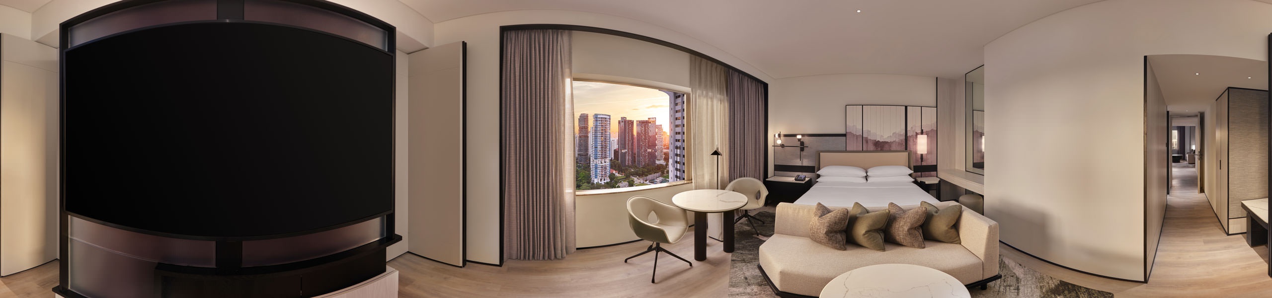 Panoramic hotel suite with a king bed, curved sofa with cushions, round table and chair, large window showing a city skyline at sunset, and a dark TV wall.