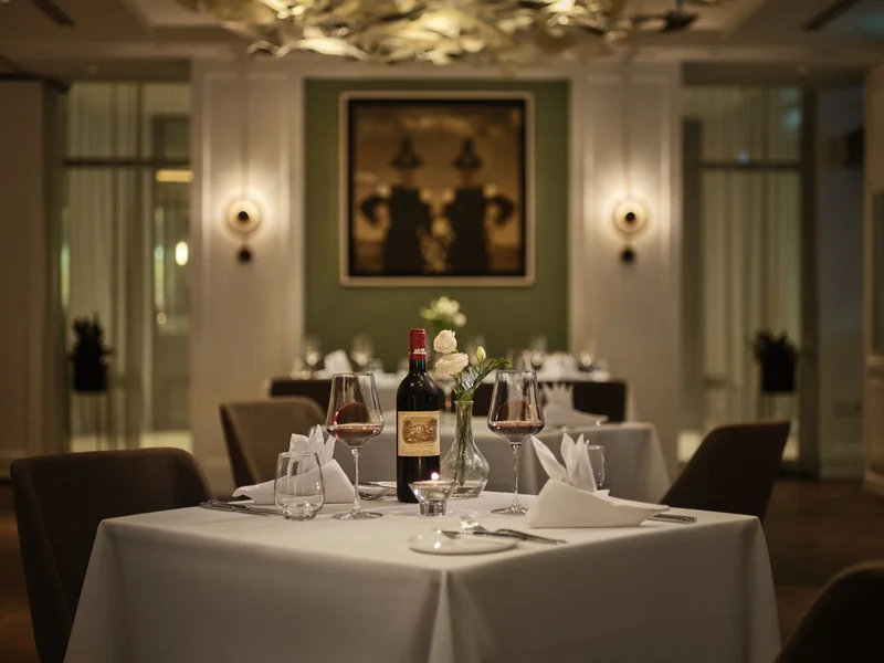 White-clothed dining table set with a red wine bottle, two filled wine glasses, white roses in a vase, and folded napkins under warm wall-sconce lighting.