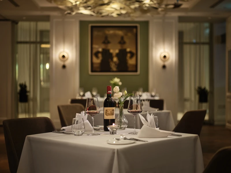 White-clothed dining table set with a red wine bottle, two filled wine glasses, white roses in a vase, and folded napkins under warm wall-sconce lighting.
