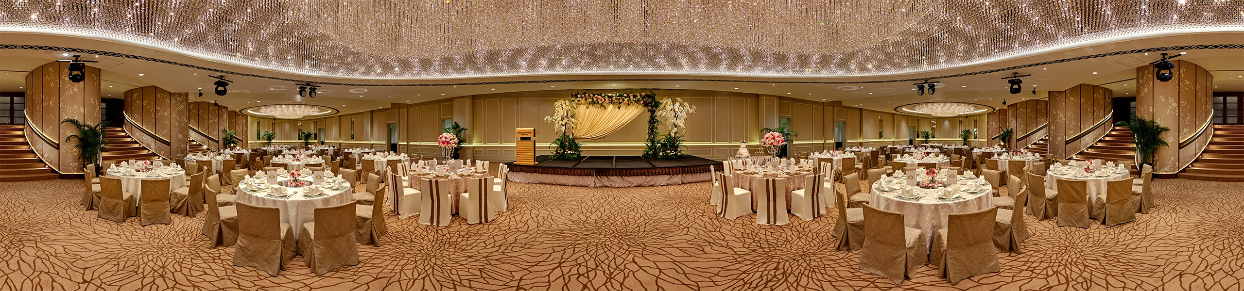 Panoramic view of a grand ballroom with a crystal chandelier ceiling, gold-draped round banquet tables, a floral stage backdrop, and a sweeping staircase.