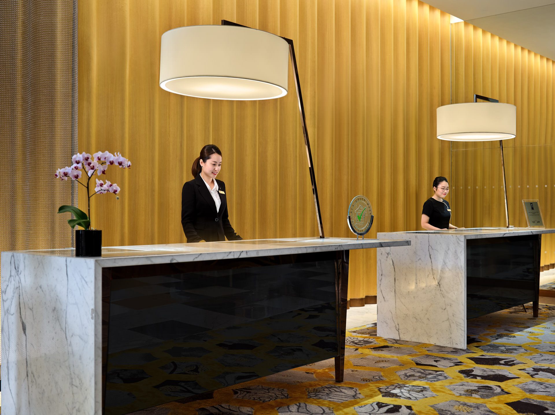 Two hotel staff in black uniforms standing behind white marble reception desks with large drum pendant lamps and a gold textured wall behind them.