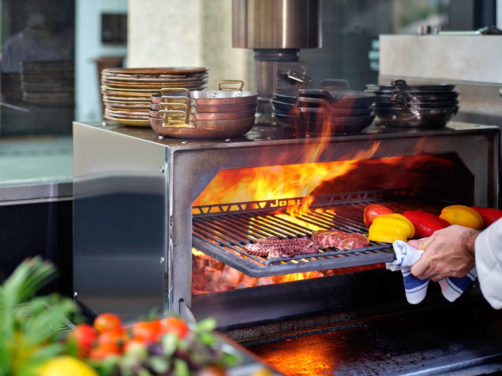 Chef grilling steak and bell peppers over open flames in a Josper charcoal oven, with copper pans and stacked plates on the shelf above.