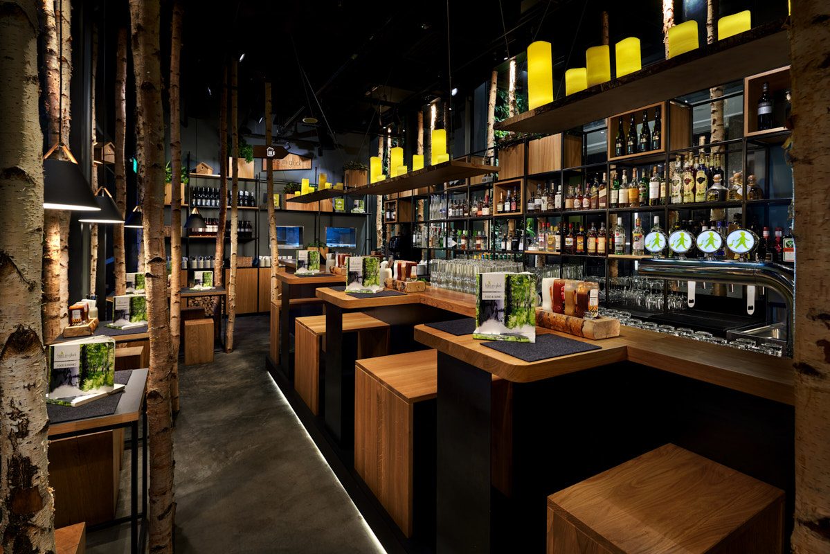 Restaurant interior with birch tree trunks as columns, wooden booth seating along a bar lined with bottles, and yellow pendant lights overhead.