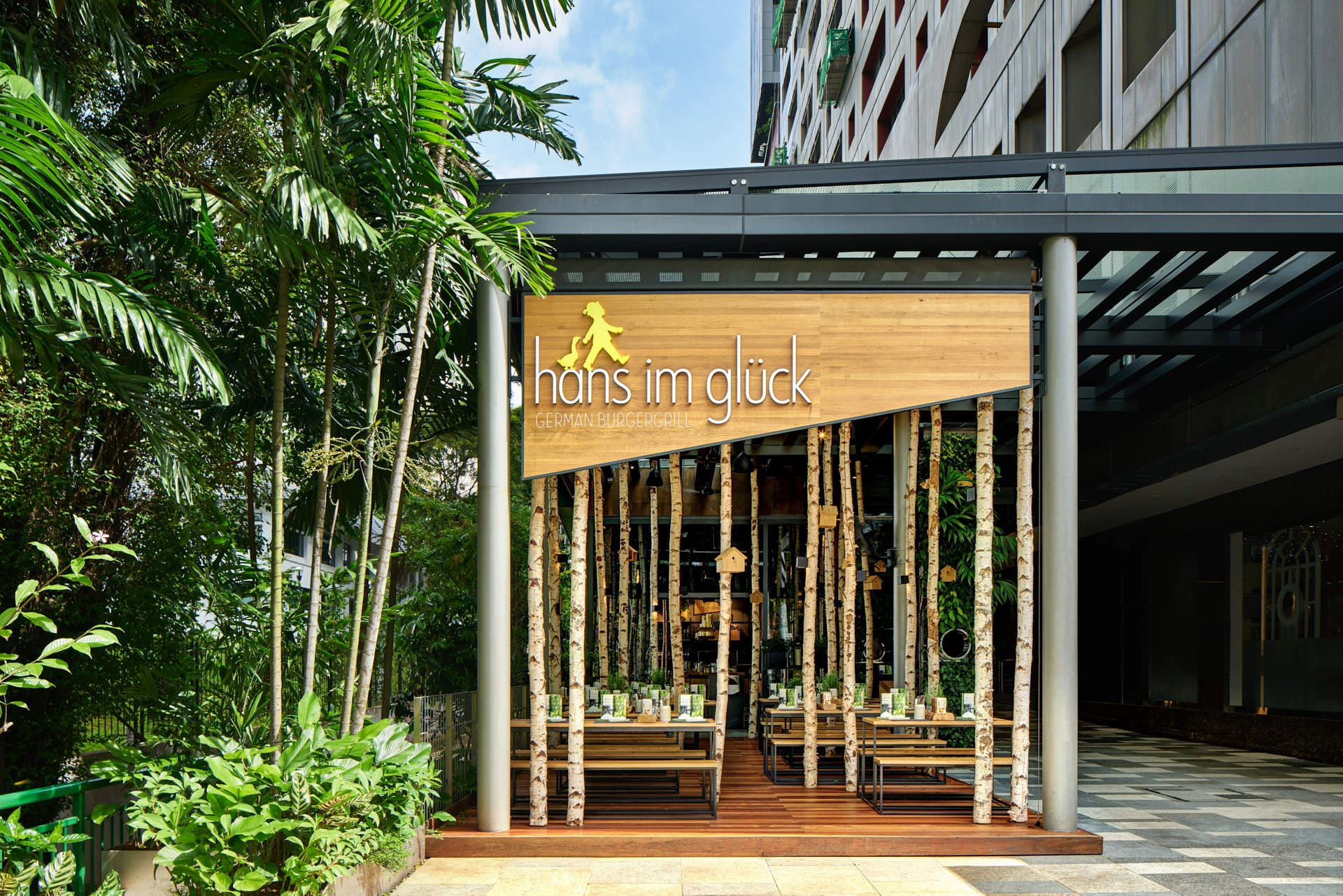Restaurant storefront with a wooden sign reading Hans im Gluck, birch tree trunks lining the open facade, and tropical greenery surrounding the entrance.