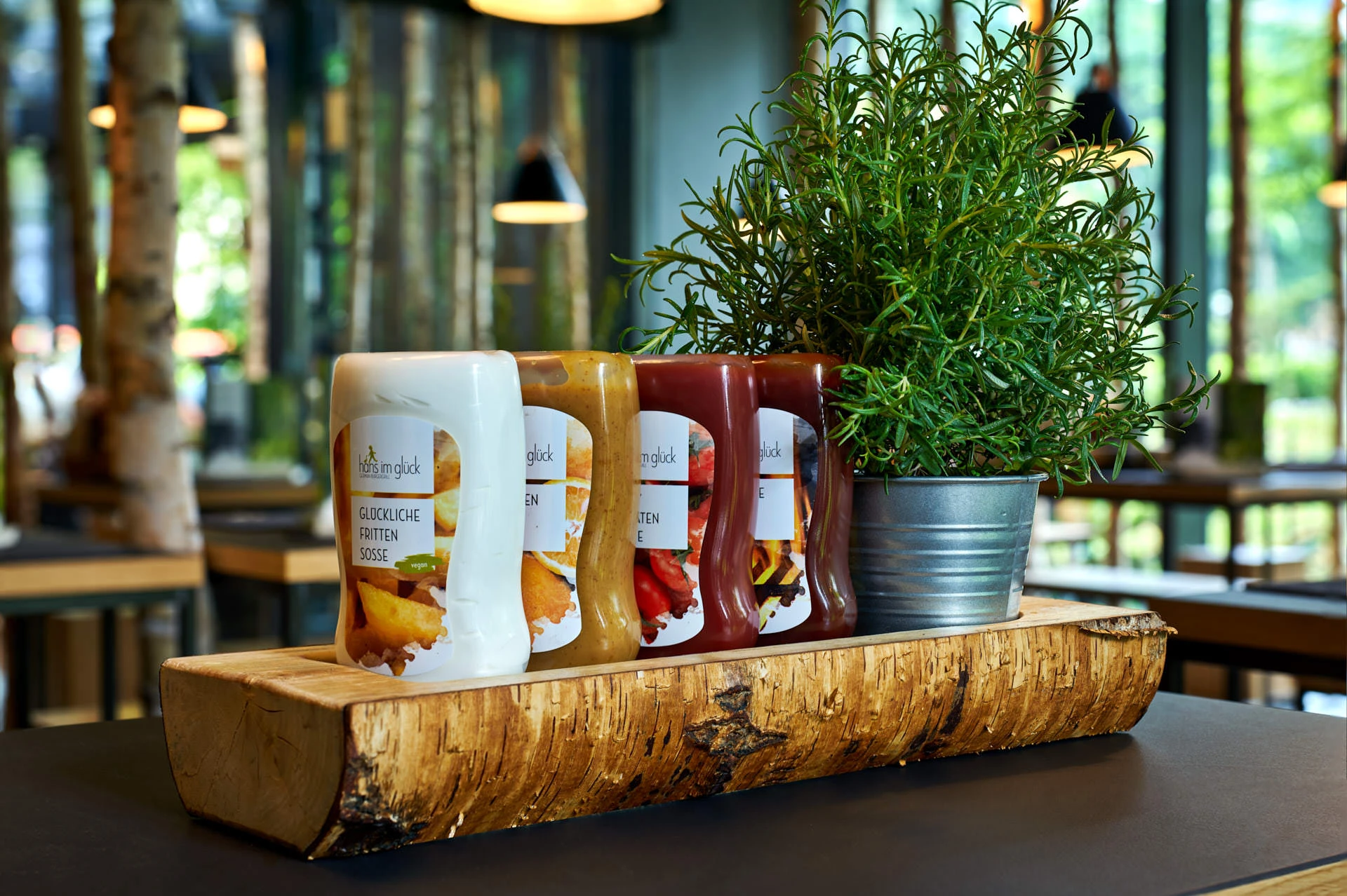 Close-up of a birch-log tray holding sauce bottles and a potted rosemary plant on a restaurant table, with blurred greenery in the background.