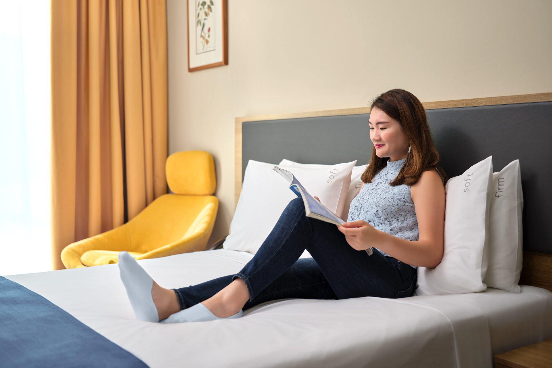 Woman reading a magazine on white hotel bed with grey headboard, yellow armchair, and amber curtains