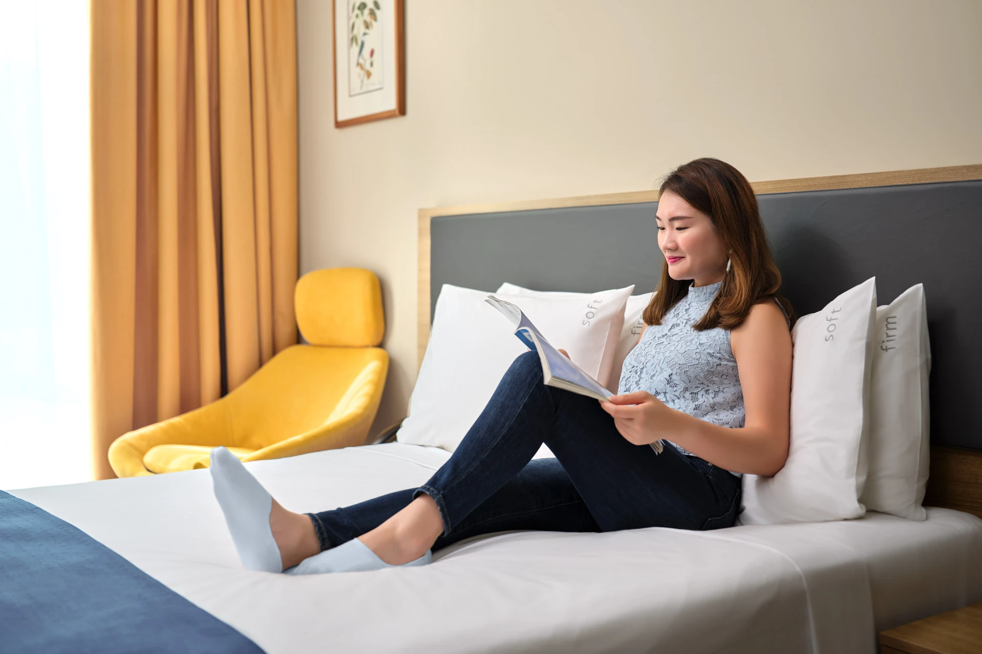 Woman reading a magazine on white hotel bed with grey headboard, yellow armchair, and amber curtains
