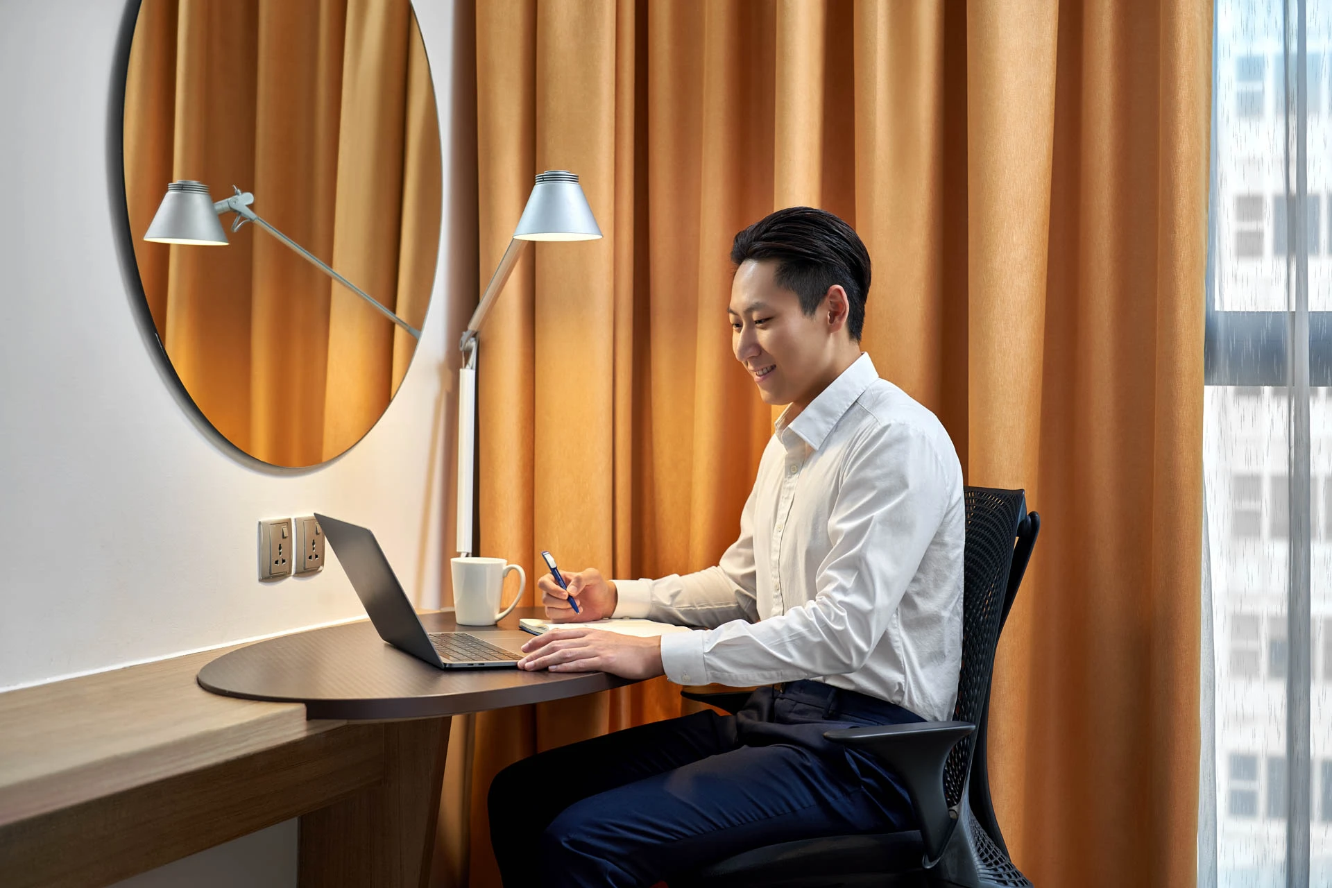 Man working at compact hotel desk with laptop, round mirror, desk lamp, and amber curtains by window
