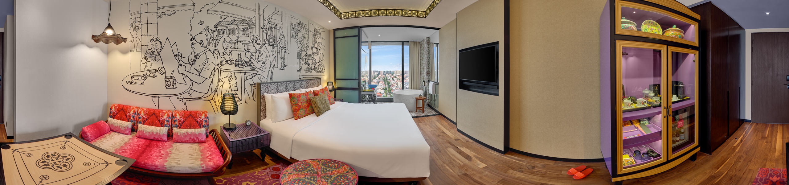 Panoramic view of a colorful boutique hotel room with white bed, red Peranakan-patterned sofa, black-and-white wall mural, and purple-lit minibar cabinet.