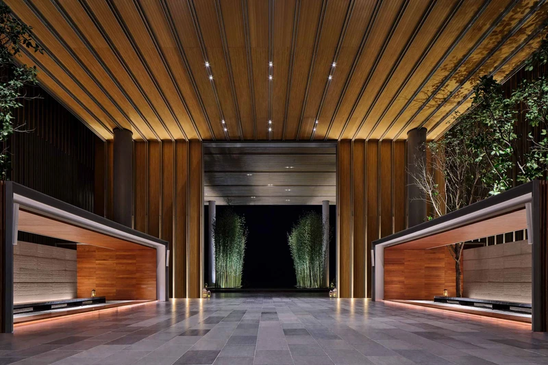Hotel entrance with slatted wooden ceiling, recessed lighting, two timber-clad reception counters, and illuminated bamboo lining the corridor beyond.