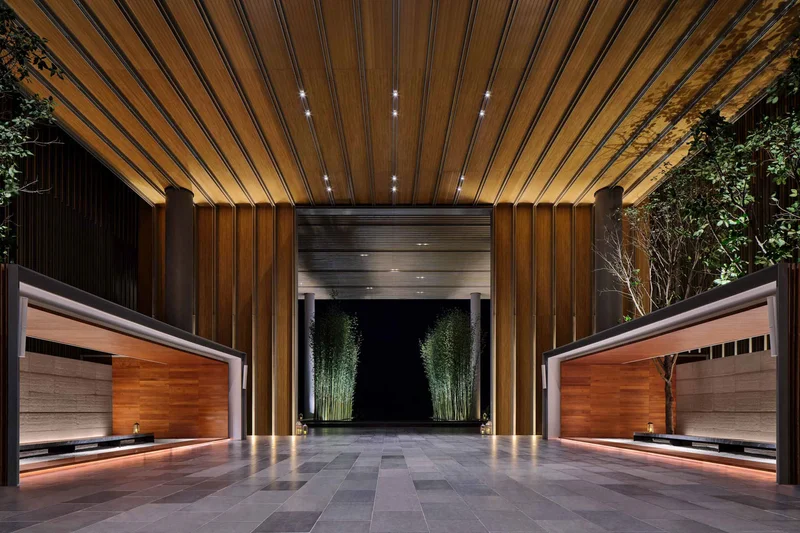 Hotel entrance with slatted wooden ceiling, recessed lighting, two timber-clad reception counters, and illuminated bamboo lining the corridor beyond.
