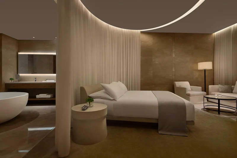 Edition Hotel Sanya suite at night with white bed, curved sheer curtain canopy, freestanding bathtub, stone-clad walls and warm ambient cove lighting