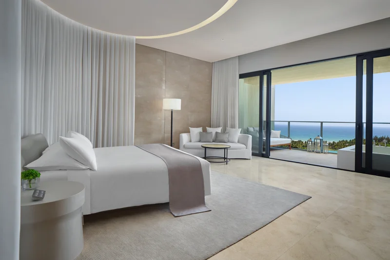 Edition Hotel Sanya suite with white bed, curved curtain canopy, lounge seating and floor-to-ceiling windows overlooking ocean and palm trees