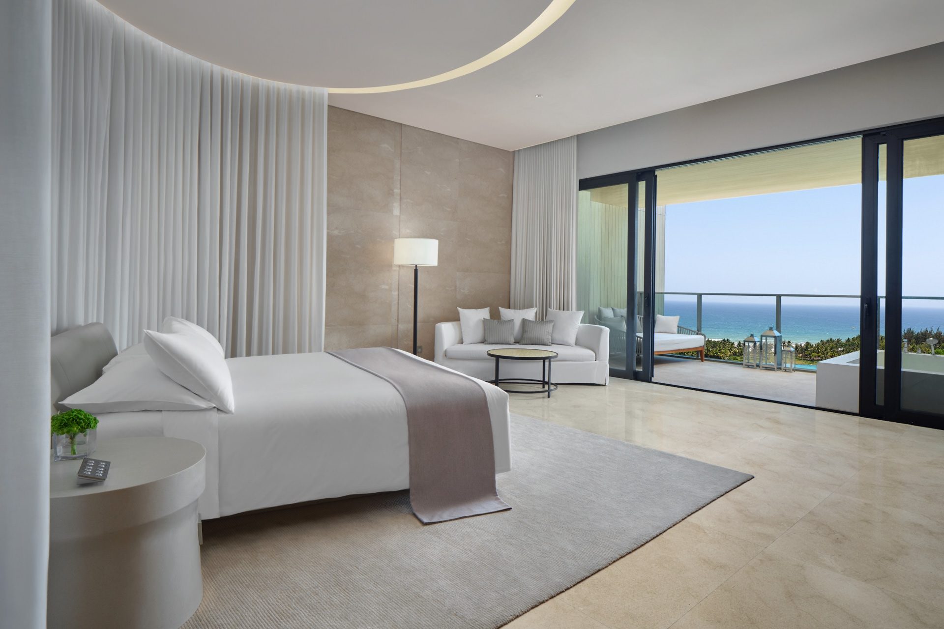 Edition Hotel Sanya suite with white bed, curved curtain canopy, lounge seating and floor-to-ceiling windows overlooking ocean and palm trees