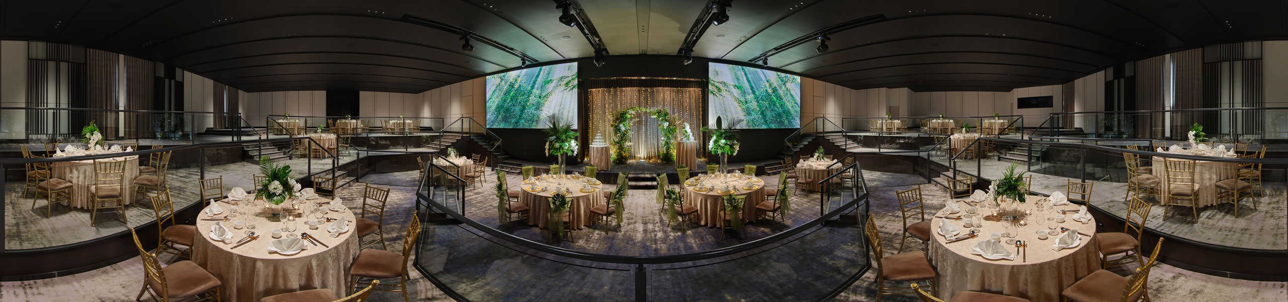 Panoramic ballroom set for a wedding with gold-clothed round tables, chiavari chairs, a greenery arch on stage flanked by two large forest-themed projection screens.