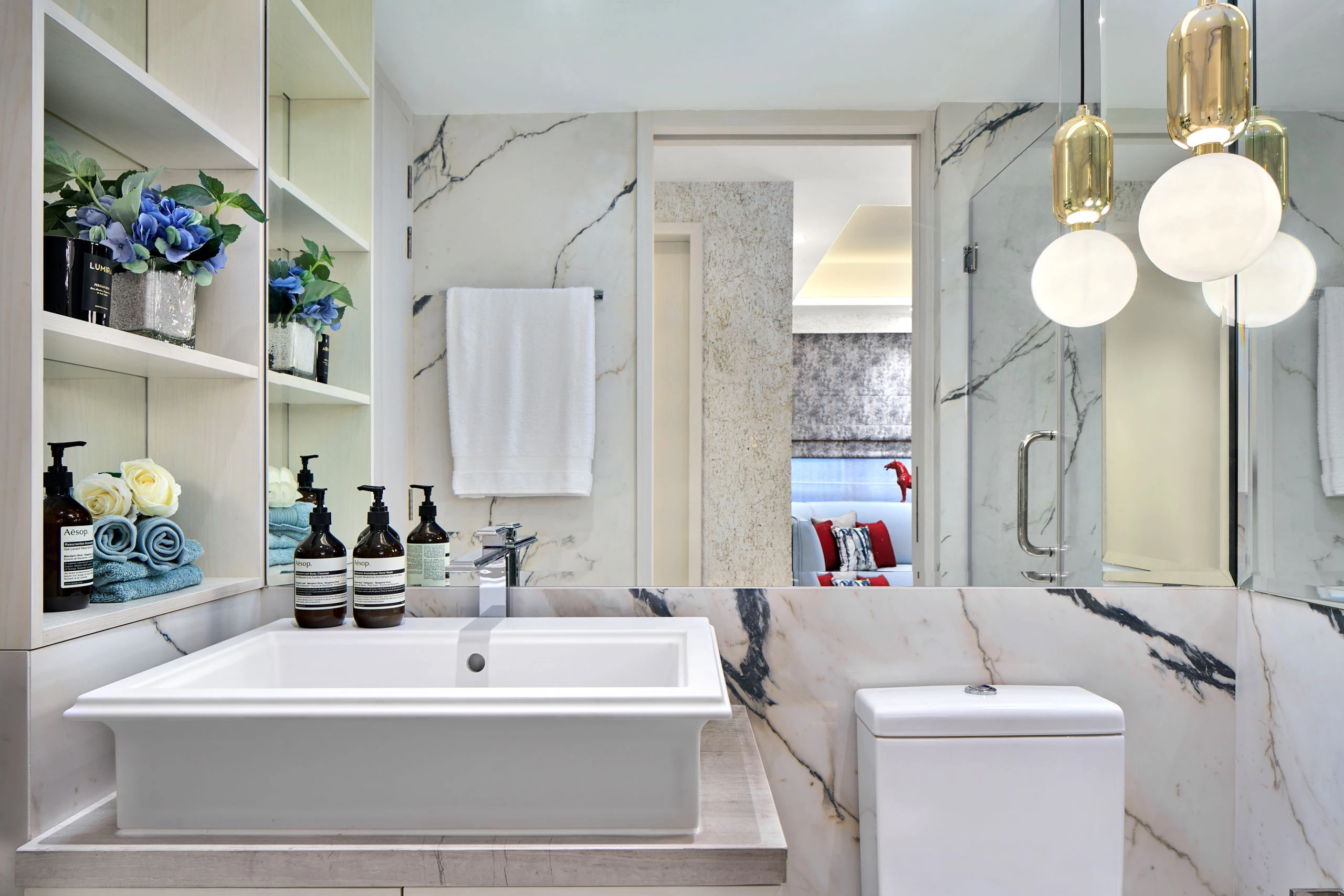 Luxury bathroom with white marble walls featuring dark veining, rectangular basin, gold pendant lights, open shelving with rolled towels and blue flowers.