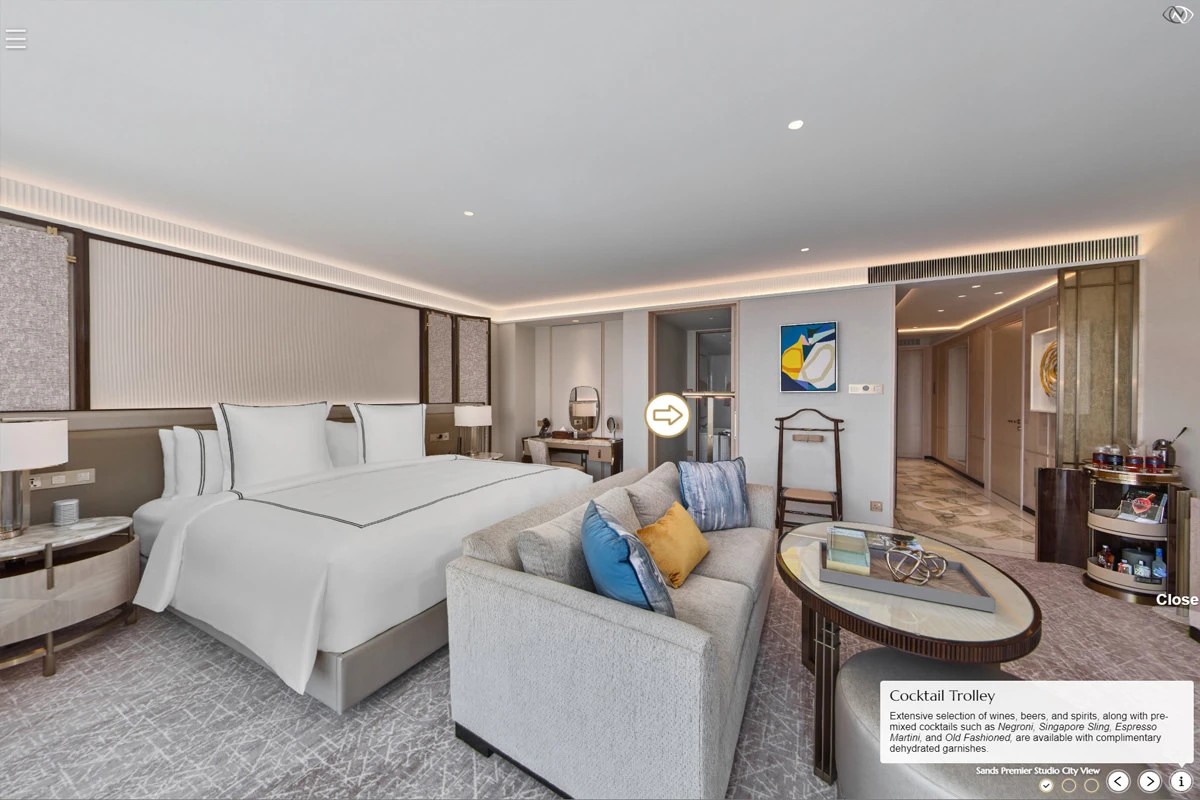Virtual tour of a spacious hotel studio room with king bed, grey sofa with blue and yellow cushions, vanity desk, valet stand, and cocktail trolley.