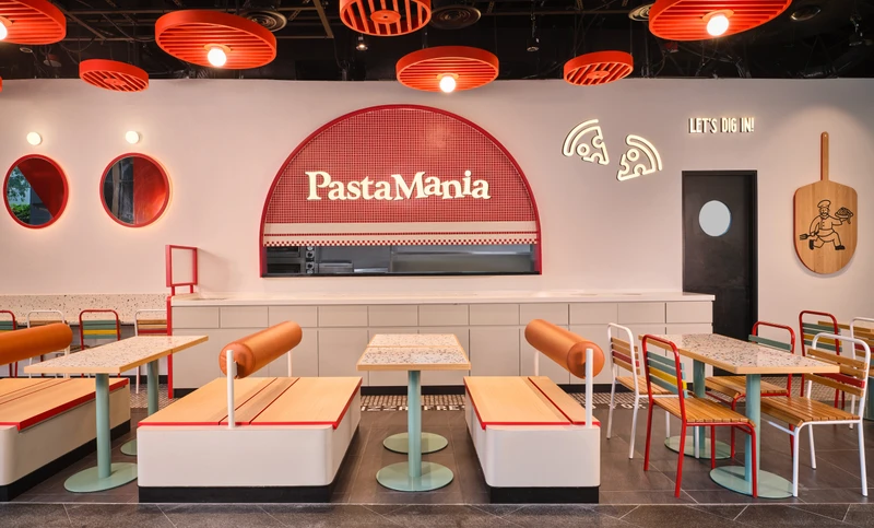 Colourful fast-casual restaurant interior with red pendant lights, booth seating, terrazzo tabletops, and a red-tiled arched service counter with PastaMania signage.