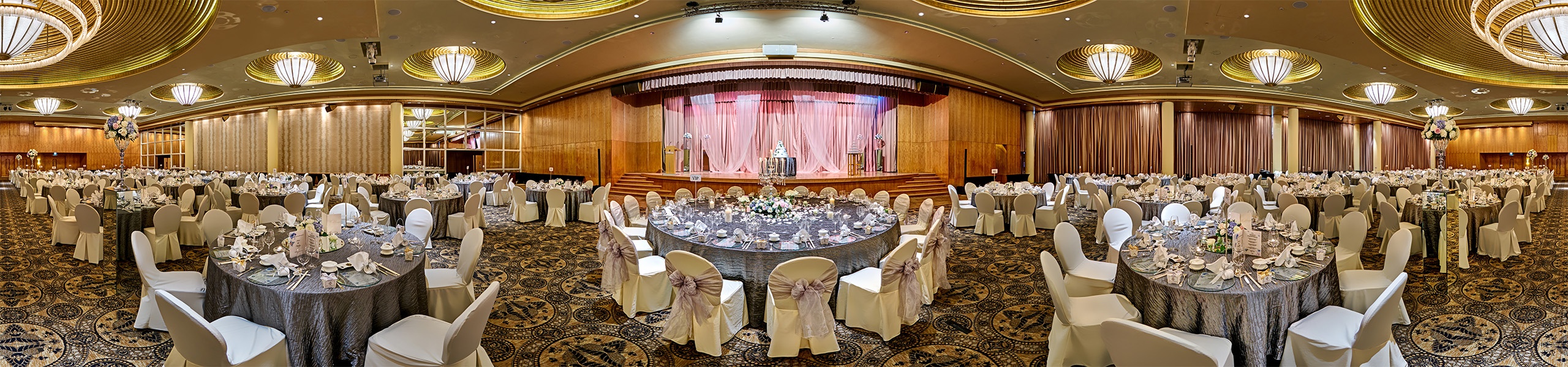 Wide panoramic view of a hotel ballroom with banquet rounds in white and grey linens, ornate patterned carpet, gold chandeliers, and a pink-curtained stage.