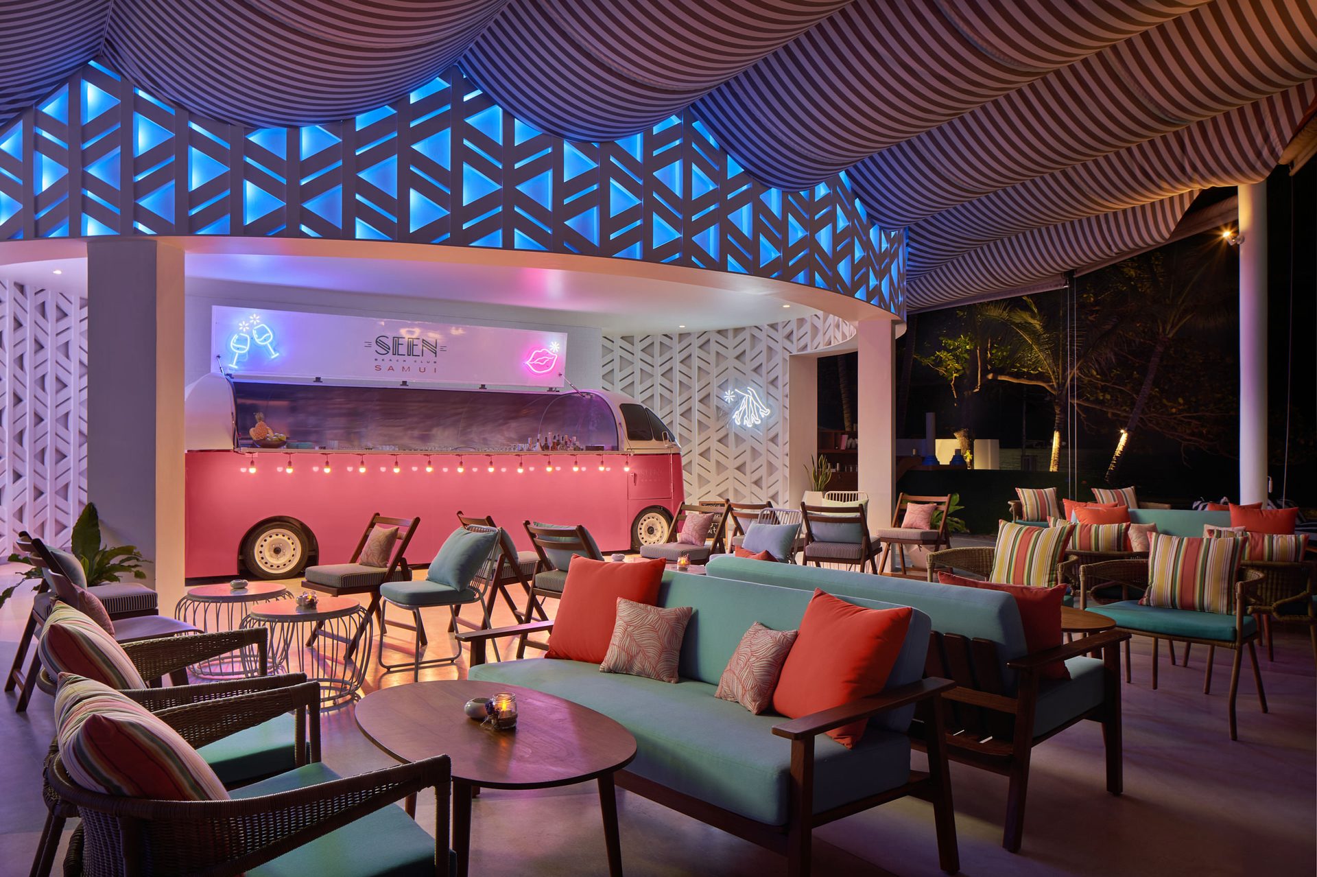 Pink vintage bus converted into a cocktail bar with neon signage under a striped canopy ceiling, blue geometric lattice screens, and teal lounge seating with colorful cushions at Seen Beach Club Koh Samui