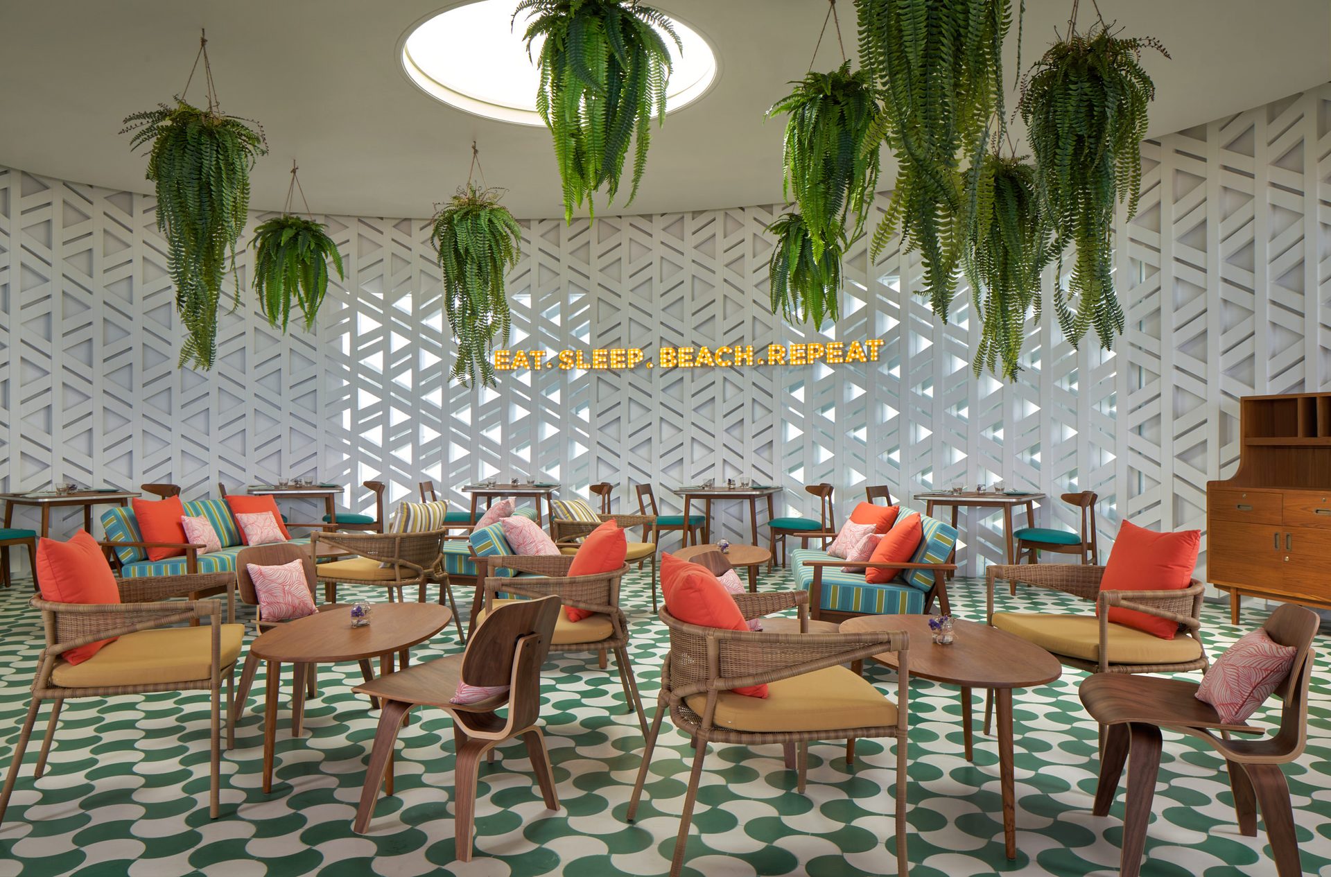 Indoor lounge with green-and-white polka-dot floor tiles, hanging ferns, white lattice walls, neon Eat Sleep Beach Repeat sign, and colorful cushioned seating under a circular skylight at Seen Beach Club Koh Samui