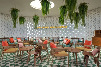 Indoor lounge with green-and-white polka-dot floor tiles, hanging ferns, white lattice walls, neon Eat Sleep Beach Repeat sign, and colorful cushioned seating under a circular skylight at Seen Beach Club Koh Samui