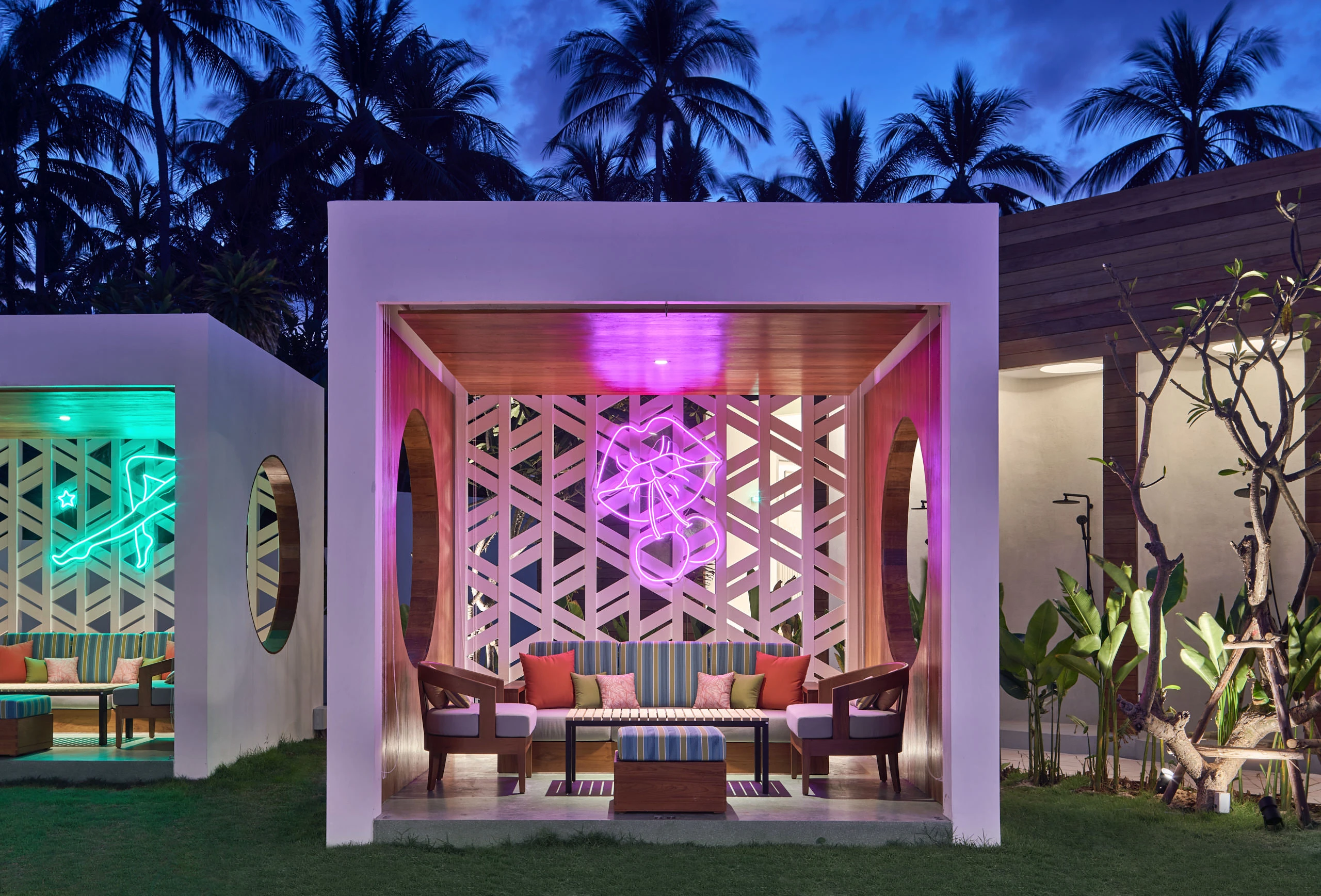 Private cabana at dusk with geometric lattice screen, pink neon artwork, striped cushioned bench seating, and palm trees silhouetted against twilight sky at Seen Beach Club Koh Samui