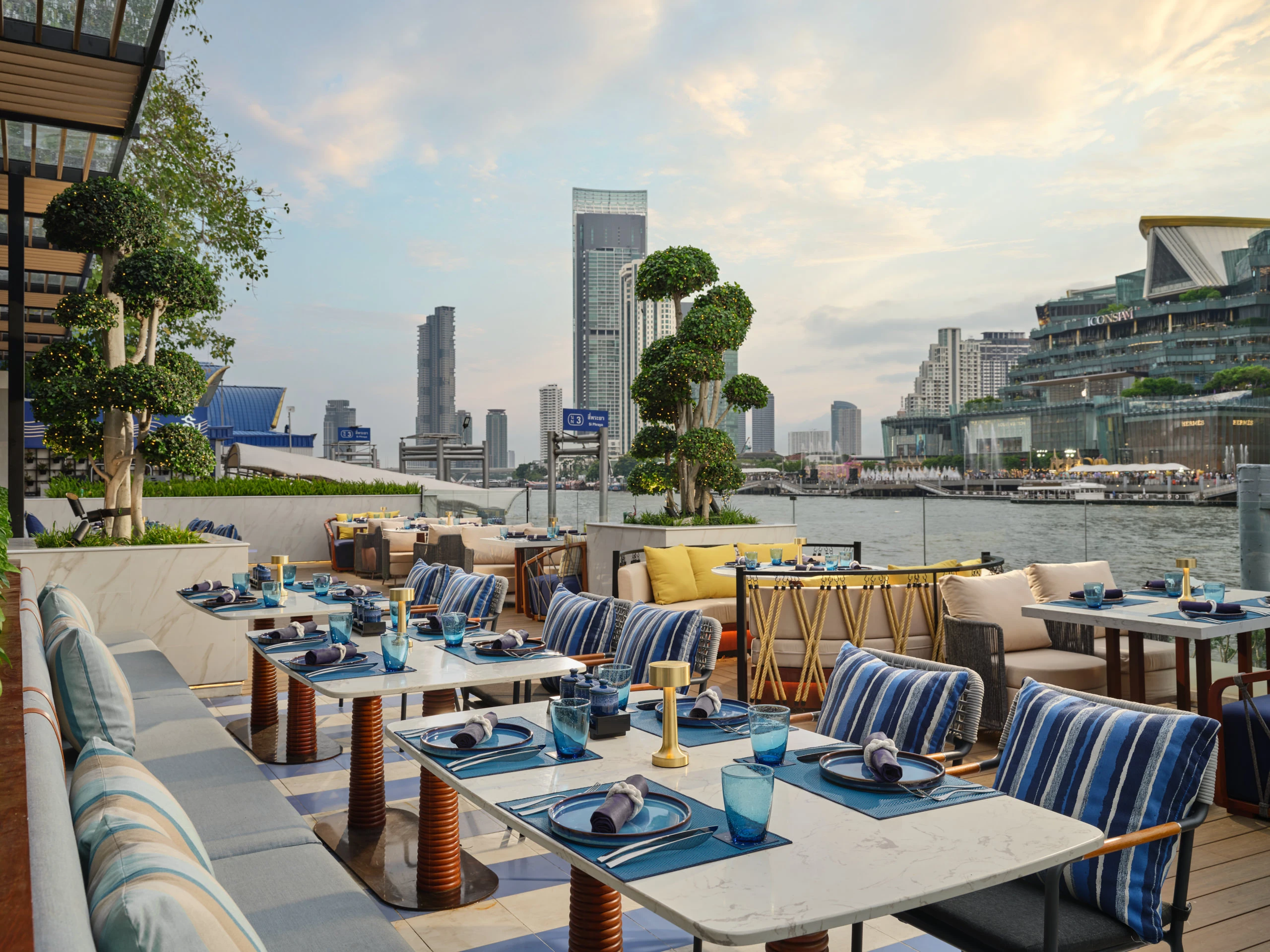 Riverfront terrace with marble tables and blue-striped cushions at golden hour — Siam Yacht Club Bangkok restaurant photography