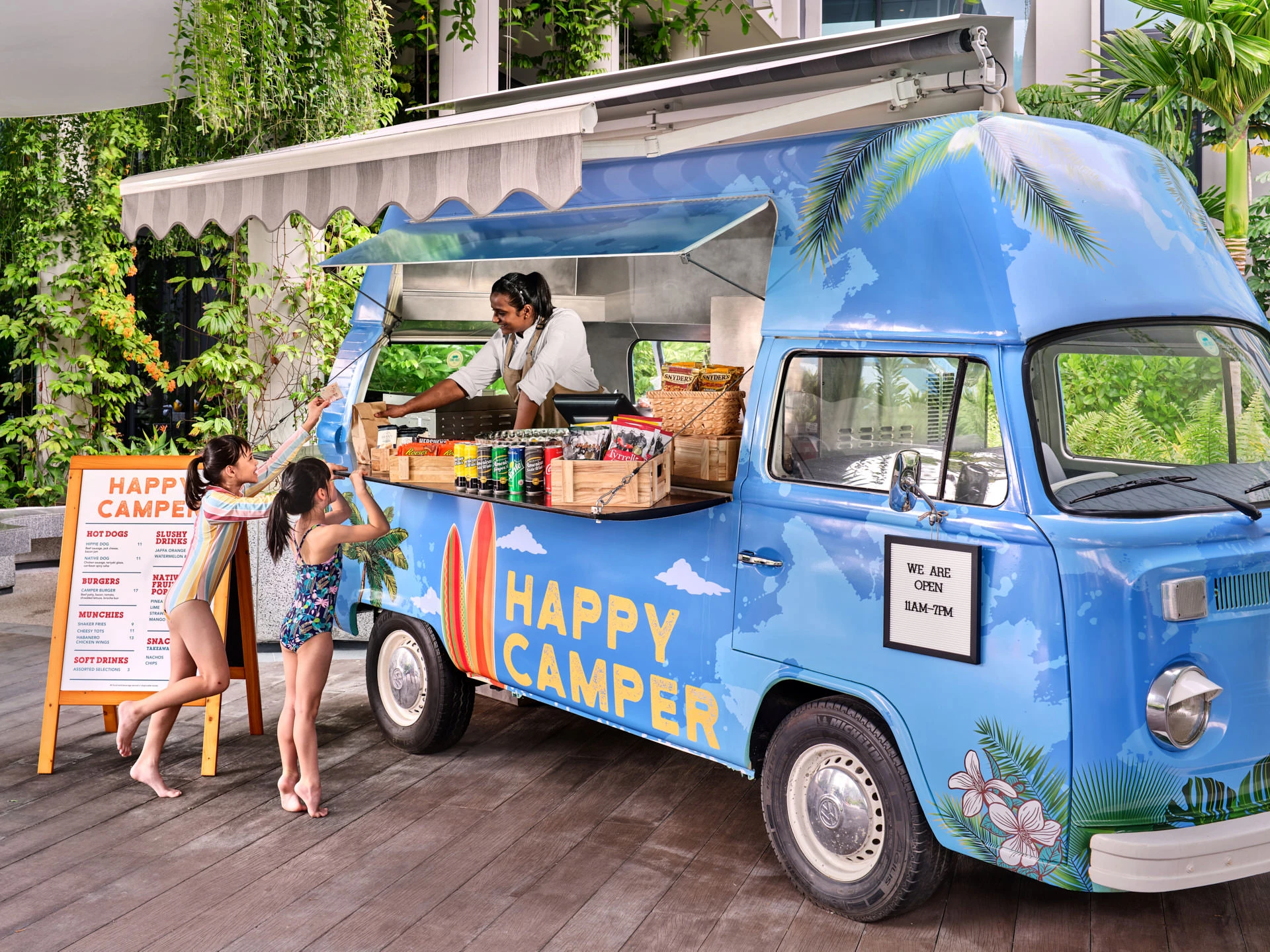 Blue vintage-style food truck labeled Happy Camper on a wooden deck, with a server handing items to two young women in swimwear.